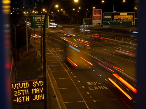 An urban highway at night with blurred motion from vehicles traveling at speed. Illuminated road signs and a digital display mentioning 'VIVID SYD' and various dates are visible. Streetlights cast a warm glow over the scene.