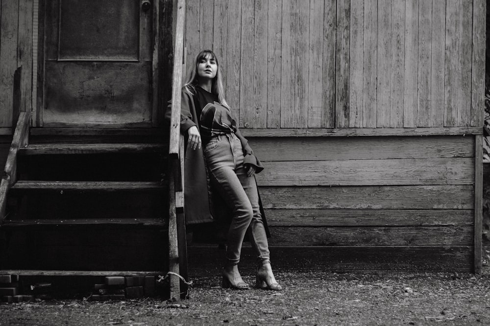 grayscale photography of woman standing and leaning against wooden house