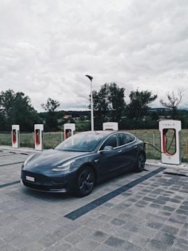 A sleek, dark-colored Tesla car is parked at a charging station with four charging units. The background features a cloudy sky and trees, suggesting a rural or semi-urban setting.