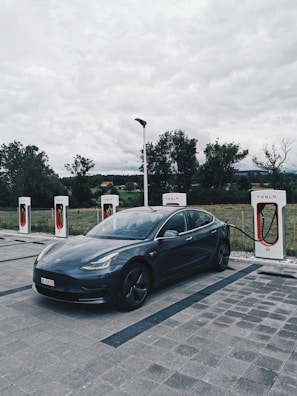 A sleek, dark-colored Tesla car is parked at a charging station with four charging units. The background features a cloudy sky and trees, suggesting a rural or semi-urban setting.