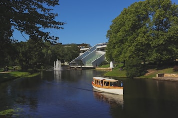 A tranquil river scene with a white and wooden boat gently floating on the water. The lush greenery of the trees lines the riverbanks, providing a serene backdrop. In the background, a modern building with large slanted glass windows is visible alongside a water fountain in the river.