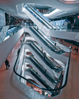 A multi-level shopping mall interior with a series of interconnected escalators. People are seen using the escalators and walking around the spacious, modern environment. The architecture features sleek lines and a combination of glass and metal elements, creating a futuristic aesthetic.