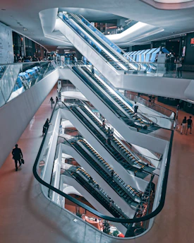 A multi-level shopping mall interior with a series of interconnected escalators. People are seen using the escalators and walking around the spacious, modern environment. The architecture features sleek lines and a combination of glass and metal elements, creating a futuristic aesthetic.