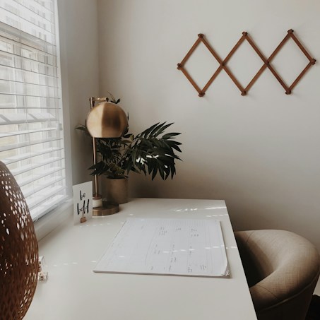 A minimalist workspace with a beige chair and a white desk. On the desk, there is a brass-colored lamp, a planner, and a pot with green leaves. A small card with the words 'be bold' is placed near the lamp. The wall behind features a decorative wooden pattern, and sunlight filters through a window with blinds.
