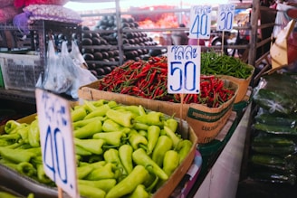 A market scene with various fresh peppers displayed in cardboard boxes. Green bell peppers, red chili peppers, and green chili peppers are prominently featured, each with handwritten price tags. The background shows more produce and market stalls.