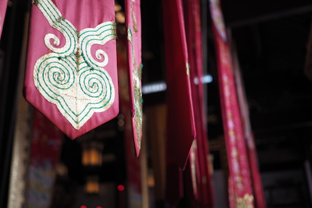 A series of vibrant red and pink fabric banners hang vertically in an indoor setting. Each banner features intricate gold and green swirling embroidery, adding a touch of elegance. The lighting is dim, with soft illumination casting shadows that enhance the rich colors and textures.