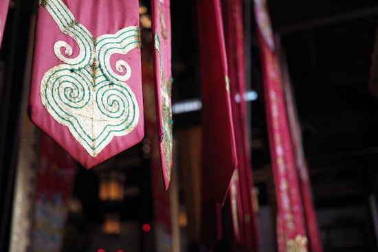 A series of vibrant red and pink fabric banners hang vertically in an indoor setting. Each banner features intricate gold and green swirling embroidery, adding a touch of elegance. The lighting is dim, with soft illumination casting shadows that enhance the rich colors and textures.