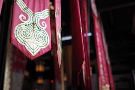 A series of vibrant red and pink fabric banners hang vertically in an indoor setting. Each banner features intricate gold and green swirling embroidery, adding a touch of elegance. The lighting is dim, with soft illumination casting shadows that enhance the rich colors and textures.