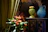 A decorative setting features a vibrant green curtain and an ornate black cabinet. On the cabinet, there is a green vase with intricate designs holding yellow and orange flowers, and a large blue ceramic pitcher containing dried wheat. Nearby, a cluster of orange anthuriums with lush green leaves adds a touch of natural beauty.