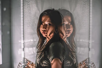 A reflective image of a woman with long hair standing against a tiled wall, creating a double exposure effect. Shadows and light patterns add depth and complexity, enhancing the mysterious atmosphere.