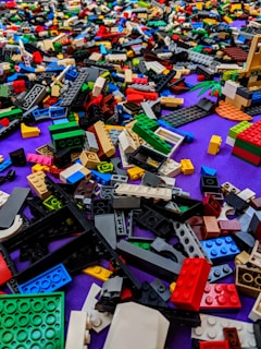 A chaotic assortment of colorful LEGO bricks scattered on a surface, featuring a variety of shapes and sizes, with a purple background visible underneath.