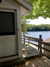 A wooden deck attached to a white building is shown, overlooking a serene lake bordered by lush green trees. The scene is shaded by an overhanging tree with bright green leaves, casting shadows on the deck and the building.