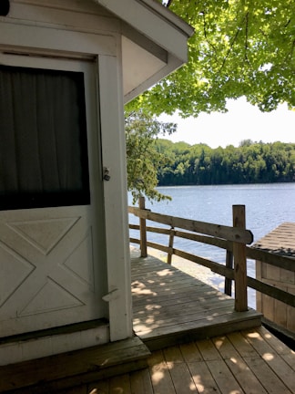 A wooden deck attached to a white building is shown, overlooking a serene lake bordered by lush green trees. The scene is shaded by an overhanging tree with bright green leaves, casting shadows on the deck and the building.