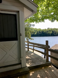 A wooden deck attached to a white building is shown, overlooking a serene lake bordered by lush green trees. The scene is shaded by an overhanging tree with bright green leaves, casting shadows on the deck and the building.