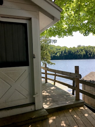 A wooden deck attached to a white building is shown, overlooking a serene lake bordered by lush green trees. The scene is shaded by an overhanging tree with bright green leaves, casting shadows on the deck and the building.
