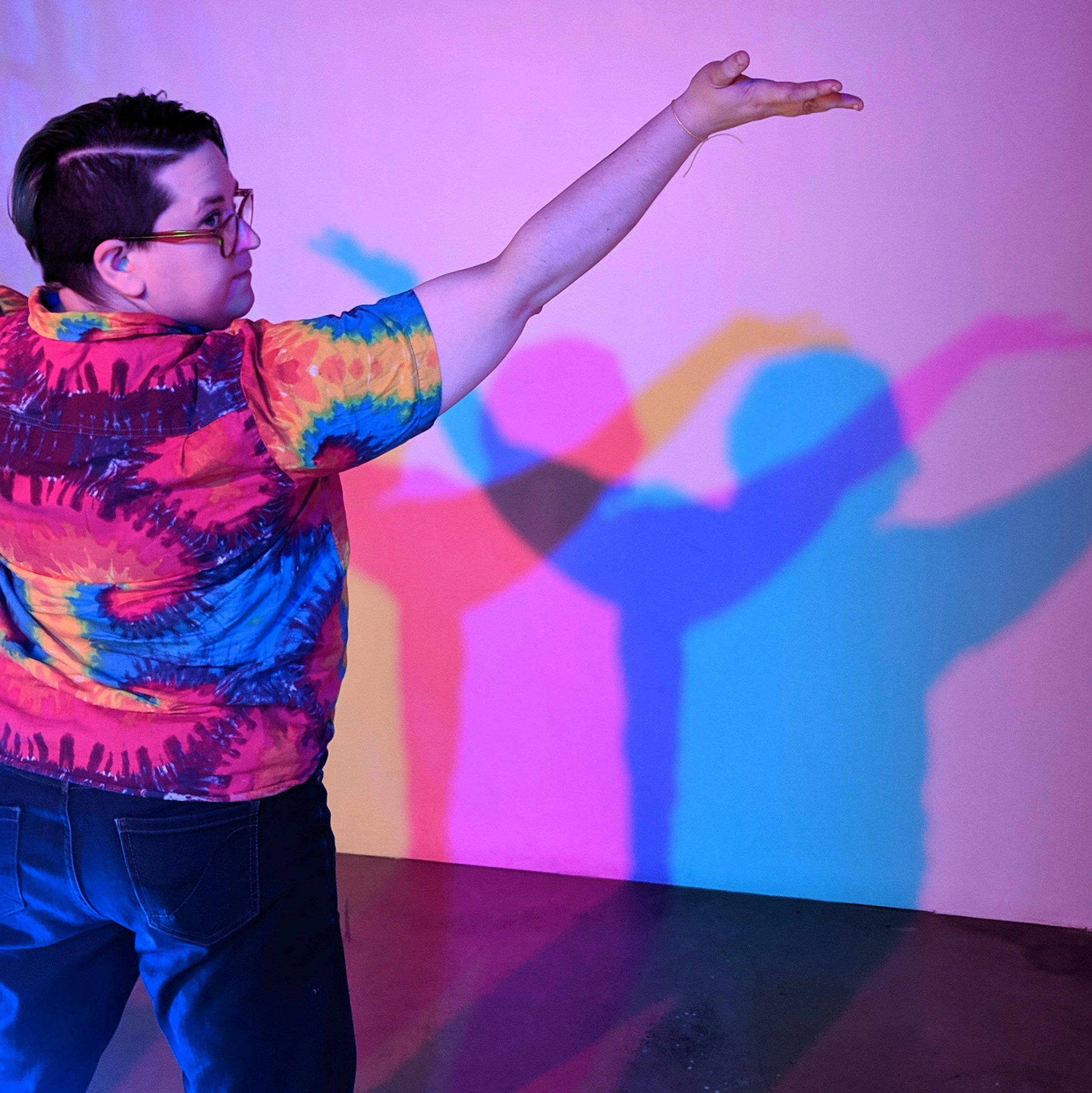 A person with short hair and glasses wearing a tie-dyed shirt strikes a pose in front of a wall, casting multi-colored shadows on it.