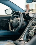 The image depicts the interior of a luxury car, focusing on the steering wheel and dashboard. The steering wheel features a prominent logo and is wrapped in black leather, with visible stitching. The dashboard displays a variety of controls and a digital screen, alongside polished materials and sleek design elements.