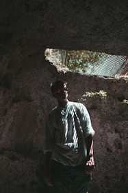 A person stands in a dimly lit cave with shadows and light patterning across their body from a narrow opening above. They are wearing a light-colored shirt and sunglasses.