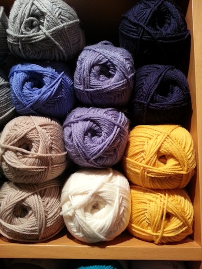 Multiple skeins of yarn neatly organized in a wooden shelf, arranged in shades of grey, blue, purple, white, and yellow. The textured fibers of the yarn provide a cozy and soft appearance.
