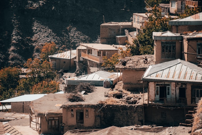 A mountainside village with rustic buildings featuring metal roofs and stone walls. The houses are closely packed together, some with wooden balconies. Trees with autumn foliage add a hint of color between the structures, and there are containers and firewood stacks visible. The village is set against a rocky, shadowed hill backdrop.