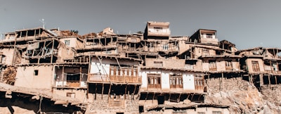 A cluster of rustic, multi-tiered houses is built closely together on a hillside, showcasing traditional architecture with a mixture of stone and wood. The buildings feature wooden balconies, weathered facades, and small windows. The overall appearance is aged, with an earthy and authentic atmosphere.