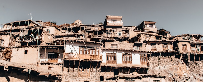 A cluster of rustic, multi-tiered houses is built closely together on a hillside, showcasing traditional architecture with a mixture of stone and wood. The buildings feature wooden balconies, weathered facades, and small windows. The overall appearance is aged, with an earthy and authentic atmosphere.