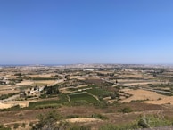 A vast rural landscape features agricultural fields with a mixture of green crops and dry land patches. In the distance, city buildings form a dense, sprawling urban area under a clear blue sky. A few scattered trees and small farmhouses can be seen across the area.