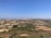 A vast rural landscape features agricultural fields with a mixture of green crops and dry land patches. In the distance, city buildings form a dense, sprawling urban area under a clear blue sky. A few scattered trees and small farmhouses can be seen across the area.