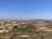 A vast rural landscape features agricultural fields with a mixture of green crops and dry land patches. In the distance, city buildings form a dense, sprawling urban area under a clear blue sky. A few scattered trees and small farmhouses can be seen across the area.