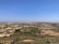 A vast rural landscape features agricultural fields with a mixture of green crops and dry land patches. In the distance, city buildings form a dense, sprawling urban area under a clear blue sky. A few scattered trees and small farmhouses can be seen across the area.