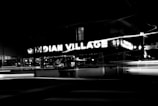 Neon lights spelling out 'INDIAN VILLAGE' are prominently displayed on a building facade, with blurred light streaks from moving vehicles in the foreground, creating a dynamic nighttime scene.
