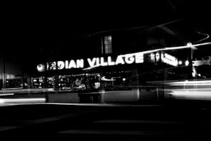 Neon lights spelling out 'INDIAN VILLAGE' are prominently displayed on a building facade, with blurred light streaks from moving vehicles in the foreground, creating a dynamic nighttime scene.