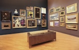 A gallery room with wooden flooring and walls displaying an eclectic collection of framed artworks, ranging from portraits to landscapes. Multiple paintings in ornate gold frames are hung on two adjacent walls, accompanied by a few sculpted busts. In the center of the room, there is a brown leather bench providing a viewing spot for visitors.