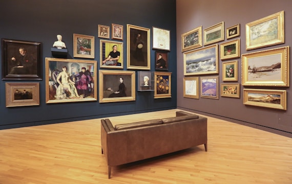 A gallery room with wooden flooring and walls displaying an eclectic collection of framed artworks, ranging from portraits to landscapes. Multiple paintings in ornate gold frames are hung on two adjacent walls, accompanied by a few sculpted busts. In the center of the room, there is a brown leather bench providing a viewing spot for visitors.