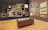 A gallery room with wooden flooring and walls displaying an eclectic collection of framed artworks, ranging from portraits to landscapes. Multiple paintings in ornate gold frames are hung on two adjacent walls, accompanied by a few sculpted busts. In the center of the room, there is a brown leather bench providing a viewing spot for visitors.