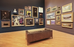 A gallery room with wooden flooring and walls displaying an eclectic collection of framed artworks, ranging from portraits to landscapes. Multiple paintings in ornate gold frames are hung on two adjacent walls, accompanied by a few sculpted busts. In the center of the room, there is a brown leather bench providing a viewing spot for visitors.