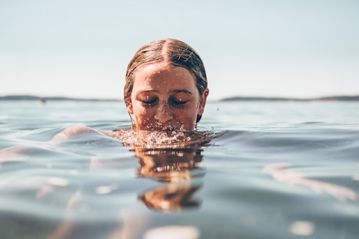 A person is partially submerged in water with their eyes closed, creating a serene and peaceful atmosphere. The water surface glistens under the sunlight, and the background reveals a distant, out-of-focus coastline.