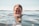 A person is partially submerged in water with their eyes closed, creating a serene and peaceful atmosphere. The water surface glistens under the sunlight, and the background reveals a distant, out-of-focus coastline.