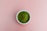 A small white bowl is filled with vibrant green powder, resting on a plain pink background.