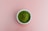 A small white bowl is filled with vibrant green powder, resting on a plain pink background.