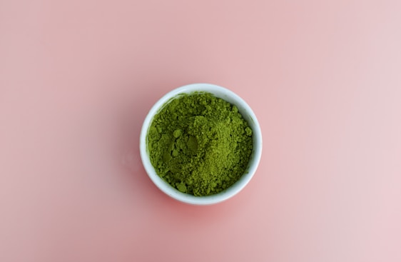 A small white bowl is filled with vibrant green powder, resting on a plain pink background.