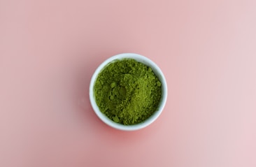 A small white bowl is filled with vibrant green powder, resting on a plain pink background.