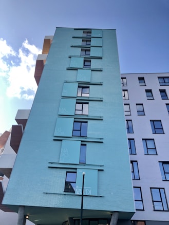 A modern multi-story building with a light blue façade featuring rectangular windows and panels with different city names. The building contrasts with the clear blue sky, which is highlighted by a few scattered clouds. The architectural design includes protruding sections and a mix of colors in other parts of the building.