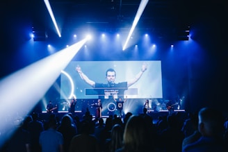 A live music performance takes place on stage with a band and a lead singer under blue and white stage lights. The audience is watching, and a large video screen displays an image of the singer with part of the lyrics.