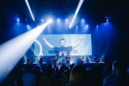 A live music performance takes place on stage with a band and a lead singer under blue and white stage lights. The audience is watching, and a large video screen displays an image of the singer with part of the lyrics.