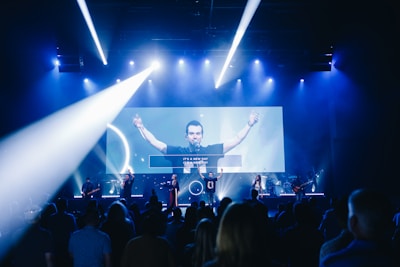 A live music performance takes place on stage with a band and a lead singer under blue and white stage lights. The audience is watching, and a large video screen displays an image of the singer with part of the lyrics.