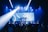 A live music performance takes place on stage with a band and a lead singer under blue and white stage lights. The audience is watching, and a large video screen displays an image of the singer with part of the lyrics.