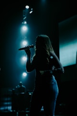 A silhouette of a person holding a microphone, likely performing on stage. Bright stage lights illuminate the background, creating a dramatic and moody atmosphere.