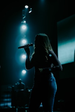 A silhouette of a person holding a microphone, likely performing on stage. Bright stage lights illuminate the background, creating a dramatic and moody atmosphere.