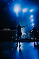 A wide shot of the band on stage with a backdrop of sound wave graphics glowing in dark blue and electric accents.